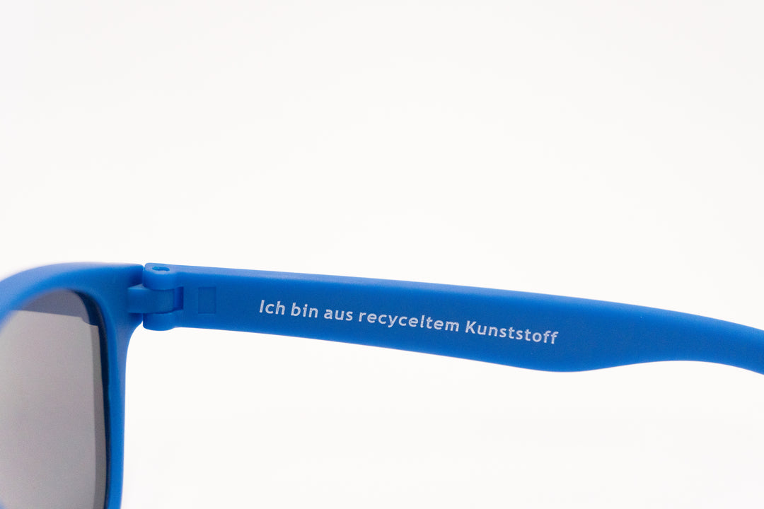Nahaufnahme des blauen Sonnenbrillenbügels mit weißem Aufdruck: „Ich bin aus recyceltem Kunststoff“.