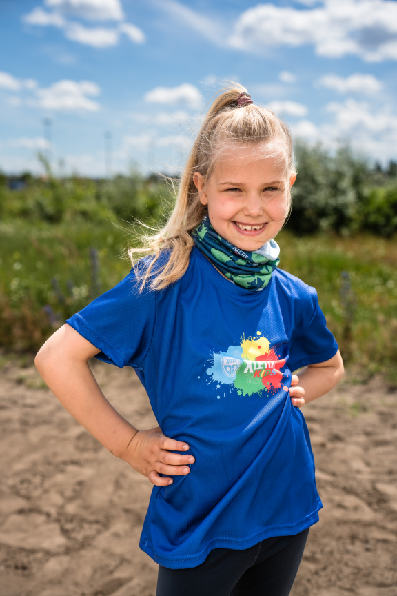 Lächelndes Mädchen auf Feld mit blauem XLETIX Kids-T-Shirt und grünem Camouflage-Multifunktionstuch.