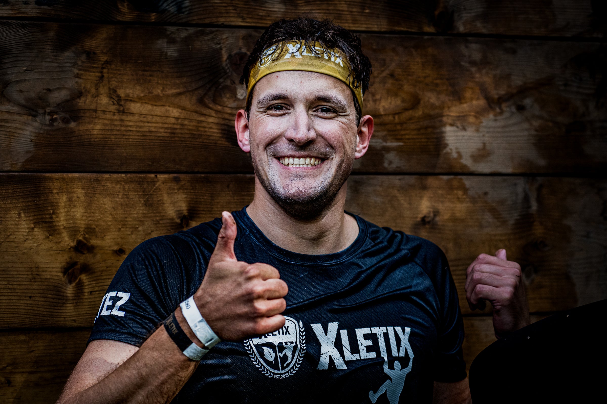 Ein lachender Mann mit XLETIX-Stirnband und Eventshirt zeigt Daumen hoch vor einer Holzplatte – sichtbar erschöpft, aber zufrieden nach dem Lauf.