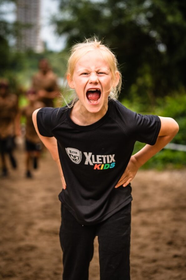 Blondes Mädchen mit offenem, schreiendem Mund in sportlicher Pose trägt ein schwarzes XLETIX Kids Funktionsshirt, Hintergrund zeigt eine Outdoor-Strecke.