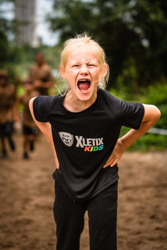 Blondes Mädchen mit offenem, schreiendem Mund in sportlicher Pose trägt ein schwarzes XLETIX Kids Funktionsshirt, Hintergrund zeigt eine Outdoor-Strecke.