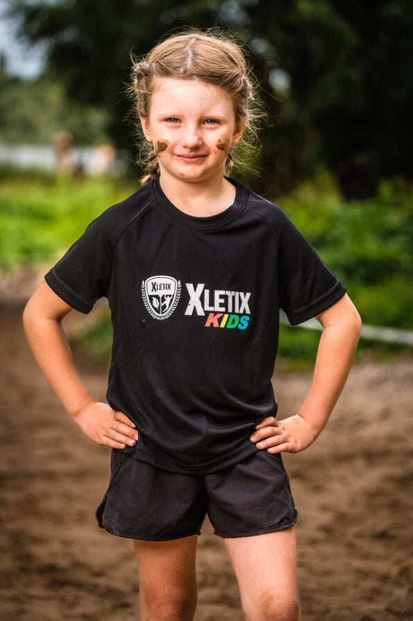Lächelndes Mädchen mit Schlammflecken im Gesicht und Händen in die Hüften gestemmt trägt ein schwarzes XLETIX Kids Funktionsshirt und Shorts.