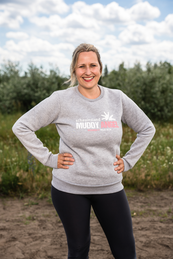 Lächelnde Frau im Freien mit grauem Muddy-Angel-Sweater und Logo-Aufdruck auf der Brust.