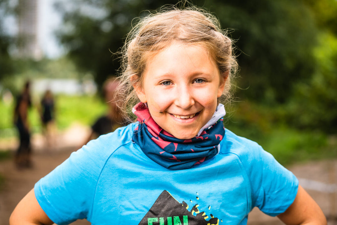 Fröhliches Mädchen mit Zöpfen trägt rot-blaues XLETIX Kids Schlauchtuch und hellblaues T-Shirt.