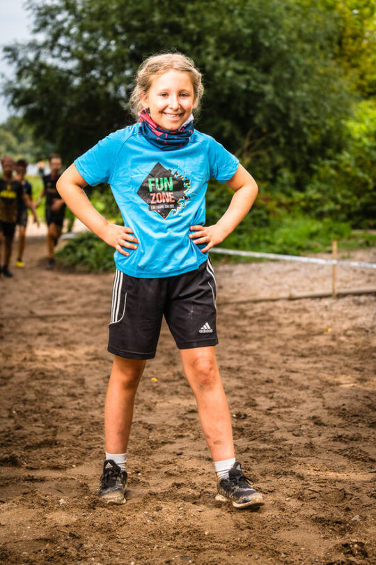 Lachendes Mädchen auf schlammigem Boden mit hellblauem „FUN ZONE“-Shirt, Schlauchtuch und Shorts.