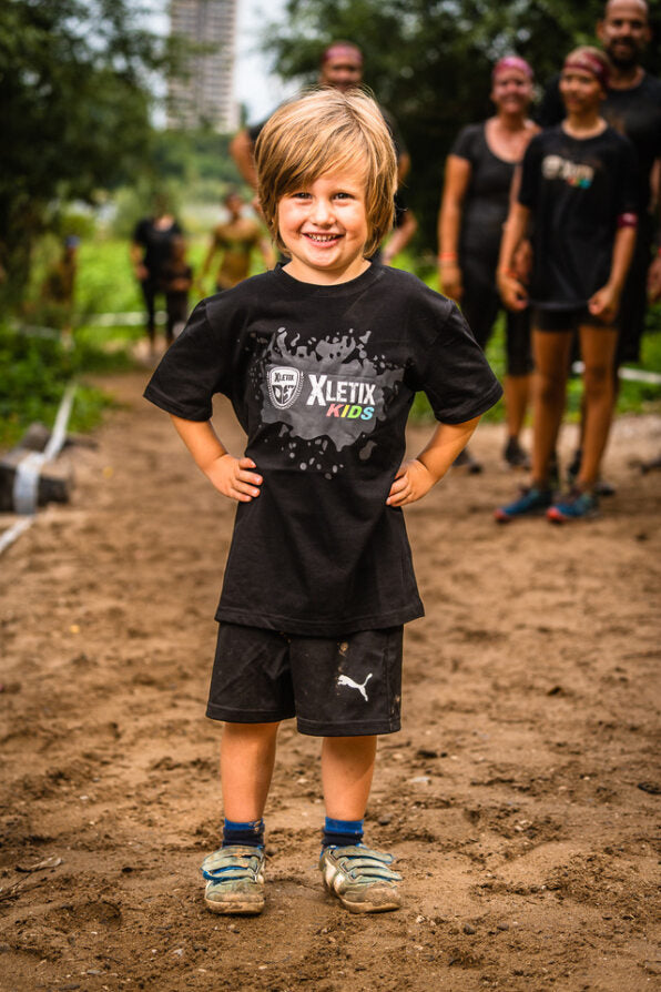 Lächelnder Junge mit blondem Haar auf matschigem Weg trägt T-Shirt mit XLETIX Kids-Logo vorn.