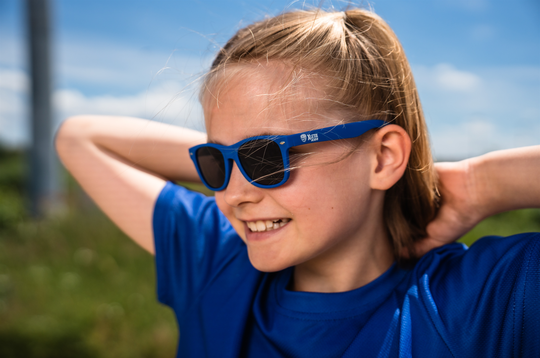 Mädchen mit blauer XLETIX Kids-Sonnenbrille und blauem Shirt lächelt und hält die Hände hinter dem Kopf.