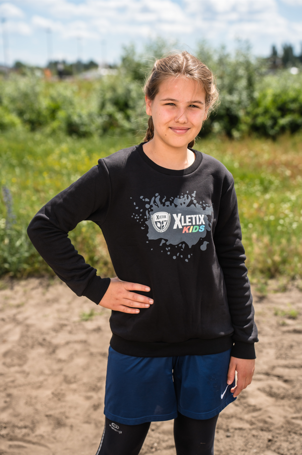 Mädchen auf Feld mit schwarzem Sweatshirt, XLETIX Kids-Logo und grauer Spritz-Grafik vorn.