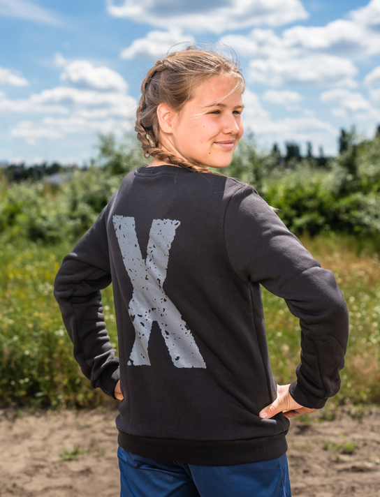 Rückseite eines Mädchens auf dem Feld mit schwarzem Sweatshirt und großem grauem „X“ auf dem Rücken.