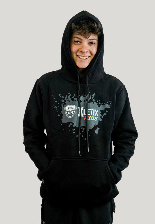 Lächelnder Jugendlicher mit schwarzem XLETIX Kids Hoodie, Farbspritzer-Print und Kapuze aufgesetzt.