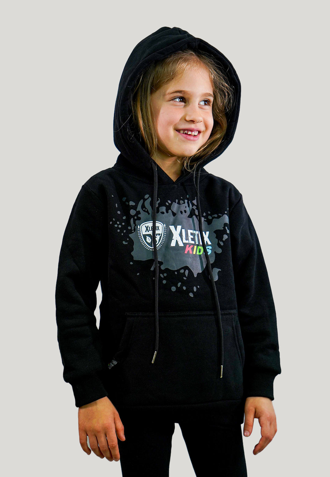 Kleines Mädchen mit schwarzem XLETIX Kids Hoodie, Farbspritzer-Design vorn und aufgesetzter Kapuze.