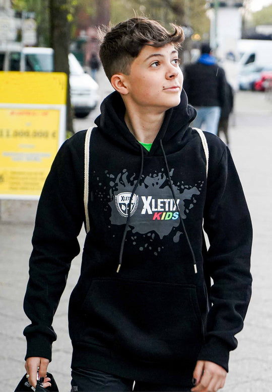 Junge mit schwarzem XLETIX Kids Hoodie läuft durch die Stadt, Kapuze auf, Blick nach oben.