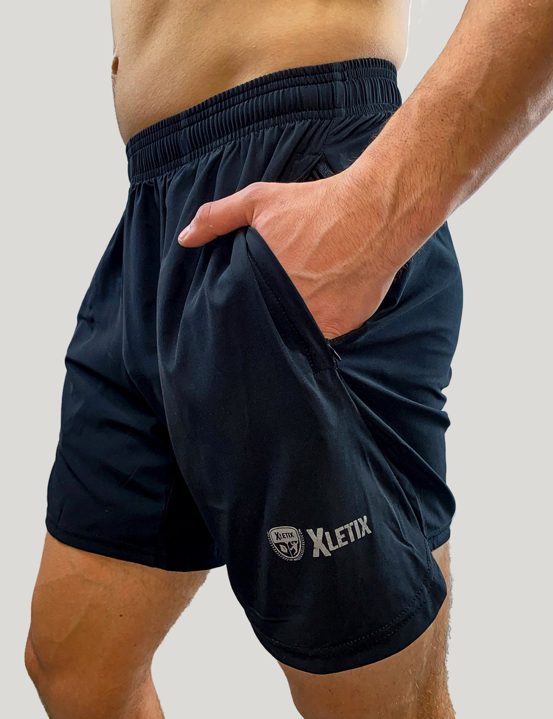 Oberkörper und Hüfte eines Mannes, er hat die schwarzen XLETIX-Shorts an, eine Hand in der Seitentasche, Logo links sichtbar.