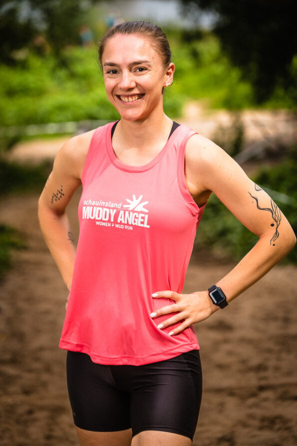 Frau trägt pinkes Muddy Angel Tanktop mit Logo, steht auf Laufstrecke und lächelt in die Kamera.
