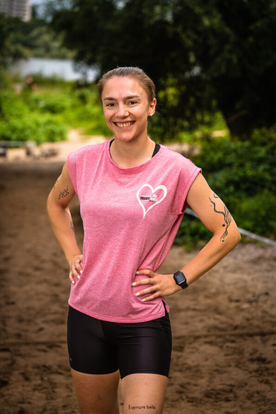 Frau trägt ein pinkfarbenes T-Shirt mit Herzlogo von Muddy Angel. Sie steht entspannt auf einem Outdoor-Parcours und lächelt.