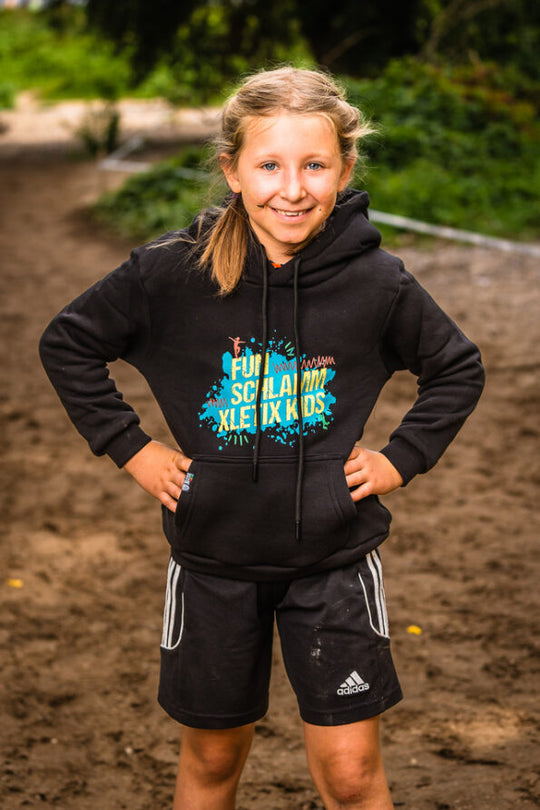 Mädchen auf sandigem Boden mit schwarzem Hoodie „FUN SCHLAMM XLETIX KIDS“, Hände in die Hüften.