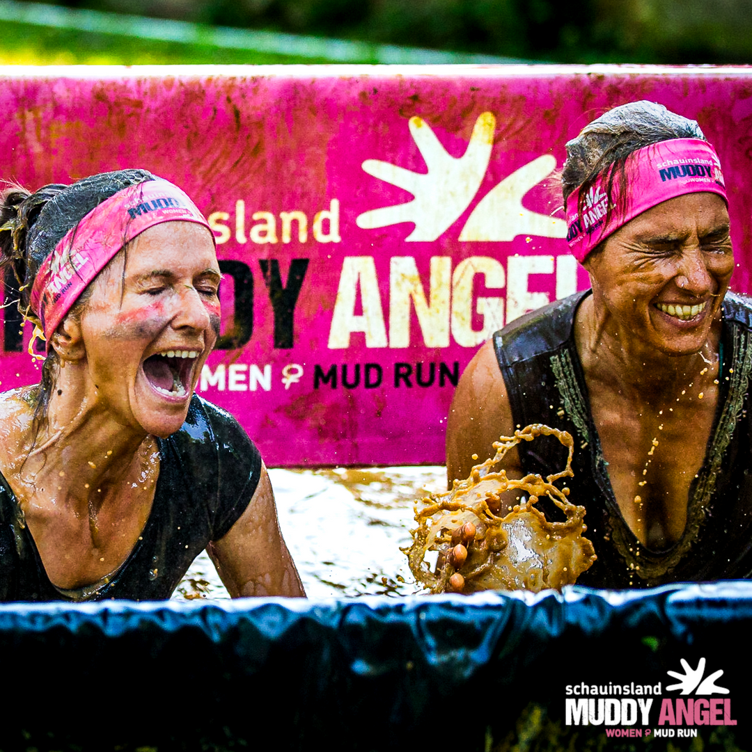 Zwei Frauen mit pinken Muddy-Angel-Stirnbändern lachen begeistert, während sie sich in einem Schlammhindernis befinden. Im Hintergrund ist ein pinkes Banner mit dem Schriftzug „Muddy Angel Women’s Mud Run“ zu sehen.