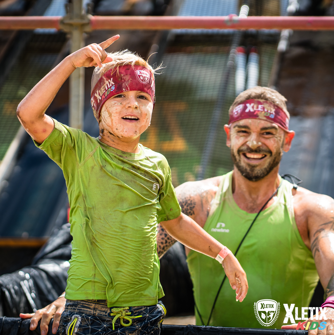 Ein schlammverschmierter Junge mit grünem Shirt und XLETIX-KIDS-Stirnband zeigt stolz in die Luft, während ein lächelnder Mann mit passendem Stirnband und Shirt hinter ihm steht. Beide wirken glücklich und sportlich aktiv. Im Vordergrund ist das XLETIX-Kids-Logo zu sehen. 