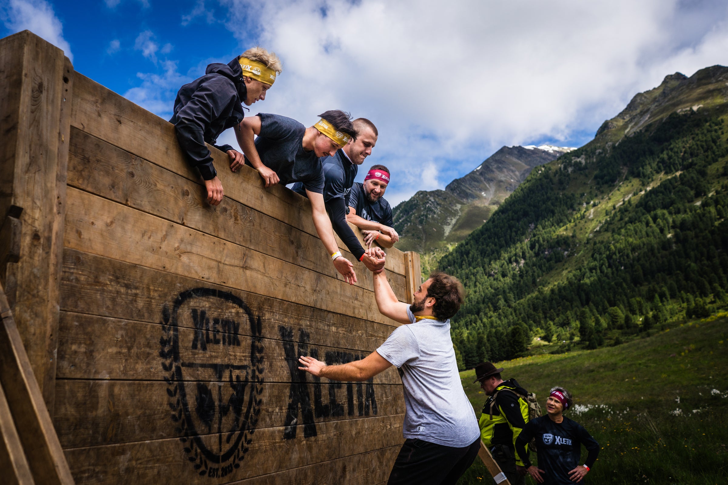 Teilnehmende eines XLETIX-Hindernislaufs helfen sich gegenseitig über eine hohe Holzwand, im Hintergrund grüne Berge und blauer Himmel. Auf der Wand ist das XLETIX-Logo sichtbar.
