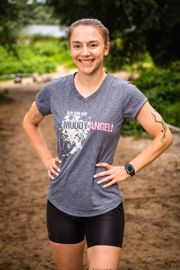 Frau im Freien trägt dunkelgraues Muddy Angel T-Shirt mit Logo und Schlammspritzer-Motiv.