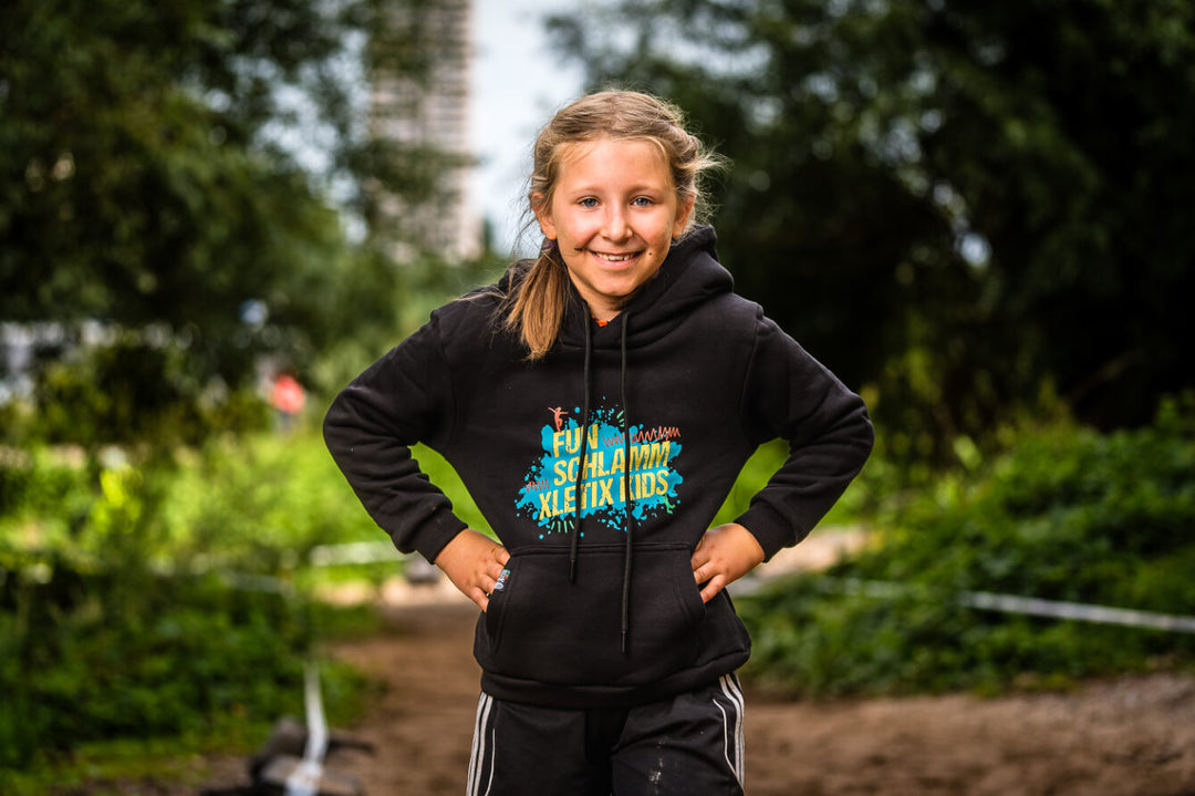 Mädchen in sportlicher Pose mit XLETIX Kids Hoodie „FUN SCHLAMM XLETIX KIDS“ vor grünem Hintergrund.