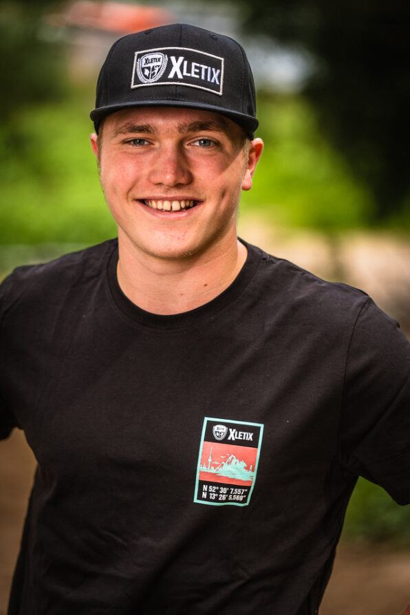 Mann mit schwarzer Snapback und Shirt mit kleinem Kartenmotiv und Koordinaten links auf der Brust.