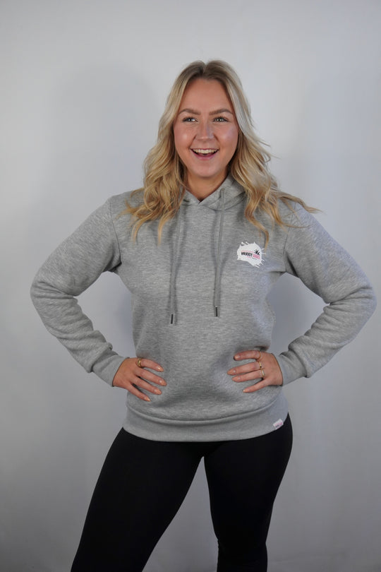 Frau trägt grauen Muddy Angel Hoodie, lächelt frontal in die Kamera, Hände in die Hüften gestemmt.